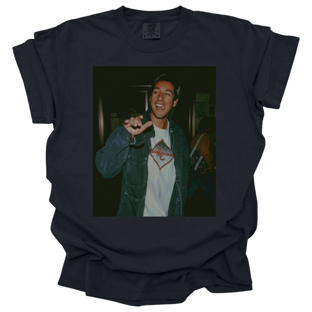 A black Comfort Colors T-shirt featuring a printed photograph of Adam Sandler waving. The vintage-style image has a nostalgic, laid-back feel, making it a statement piece for fans of classic '90s and 2000s pop culture