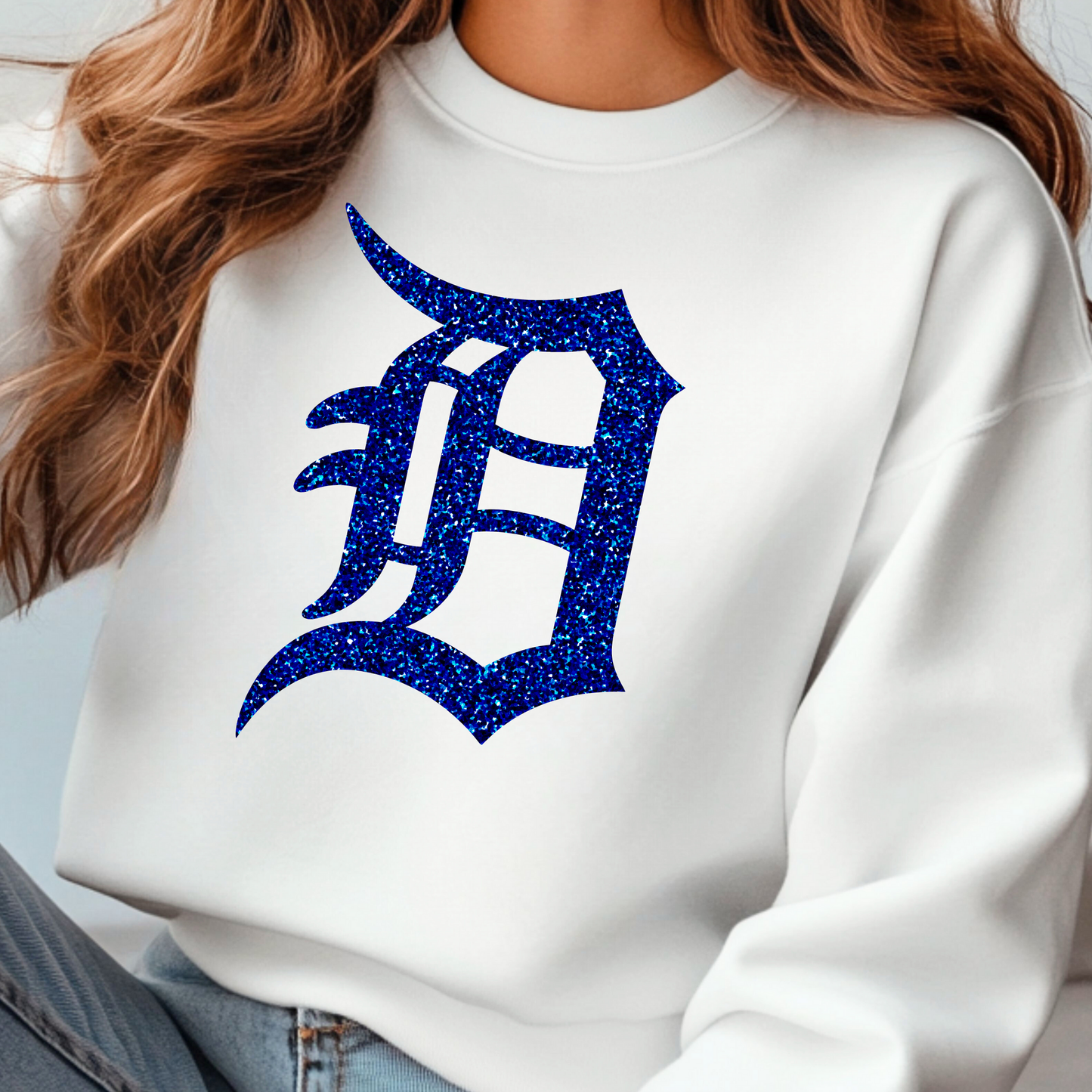 A white crewneck sweatshirt featuring a bold, glitter-textured ‘D’ in a classic Detroit-style font. The design is shown in blue glitter, adding a sparkling touch to a sporty and stylish look.