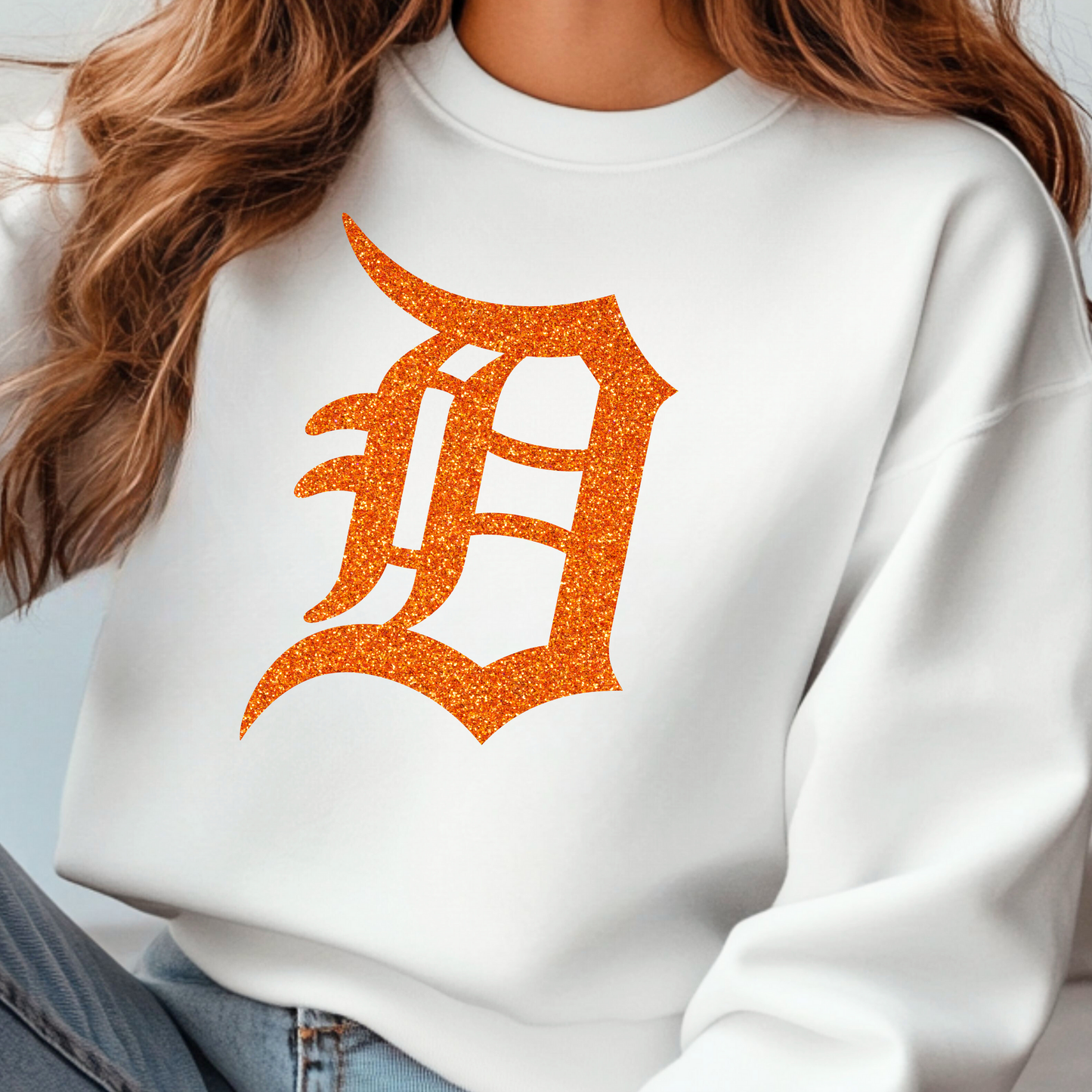 A white crewneck sweatshirt featuring a bold, glitter-textured ‘D’ in a classic Detroit-style font. The design is shown in orange glitter, adding a sparkling touch to a sporty and stylish look.