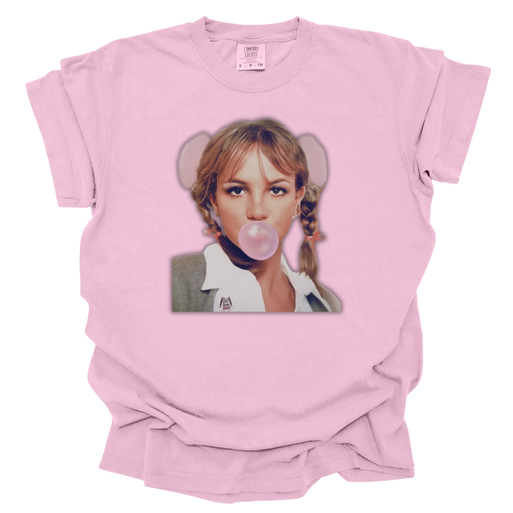 A pink Comfort Colors T-shirt featuring an image of iconic Britney Spears from her ...Baby One More Time era, blowing a pink bubblegum bubble. She is dressed in her signature schoolgirl outfit with braided pigtails, a white collared shirt, and a gray cardigan. The design captures the ultimate late '90s and early 2000s pop nostalgia.