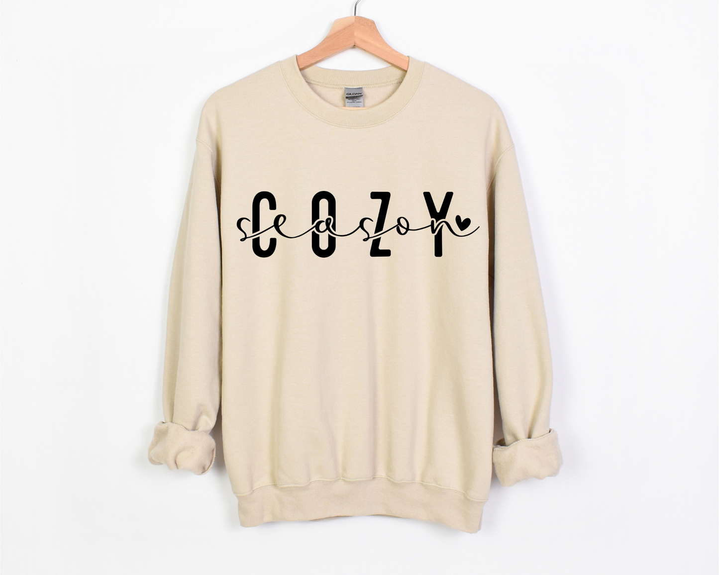 A sand-colored crewneck with the words 'Cozy Season' printed across the front. The design features bold, blocky 'COZY' lettering with 'season' written in a delicate, cursive script weaving through the letters, ending in a small heart.