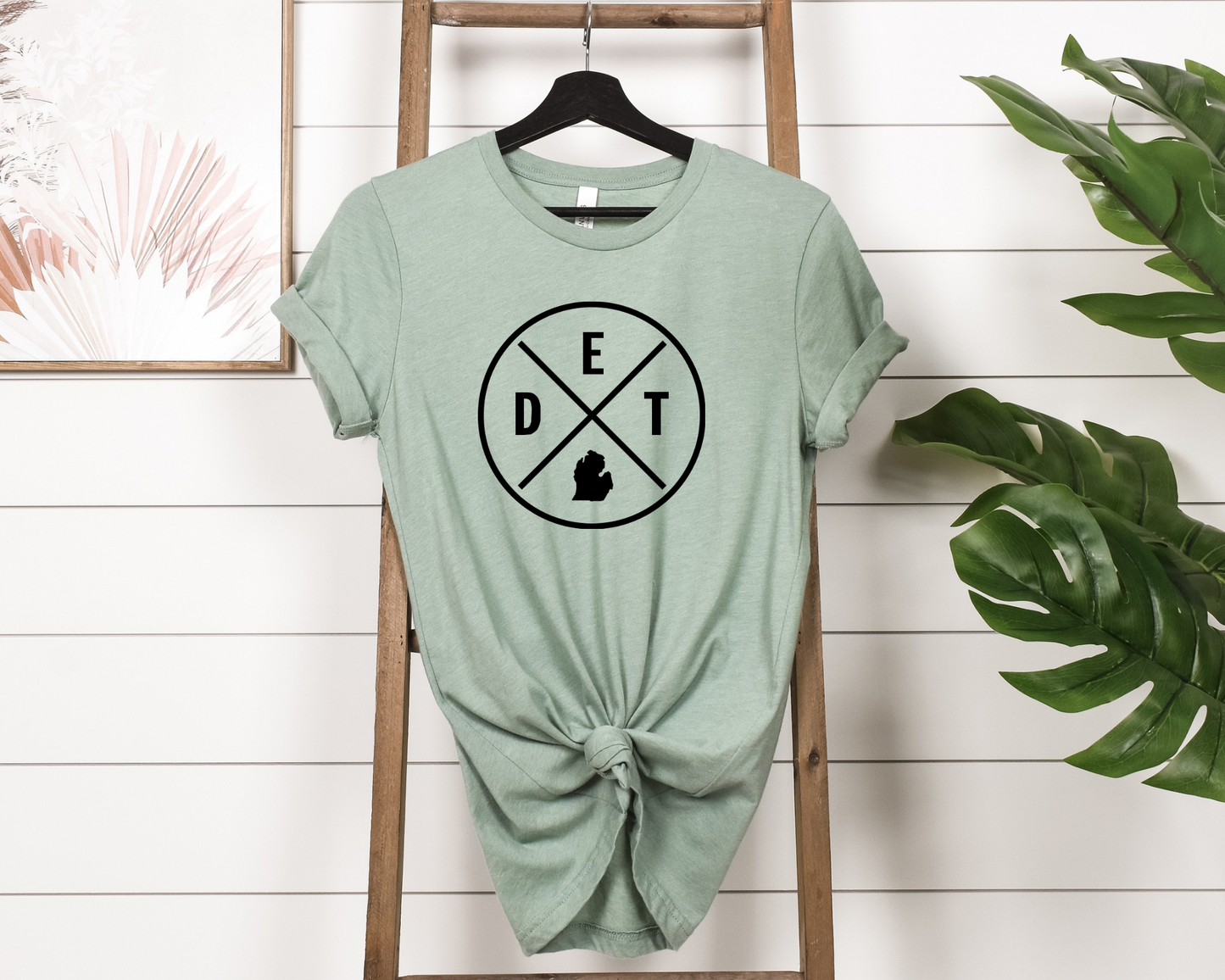 The DET MI Tee is a sage colored t-shirt that features a minimalist circular design with "DET" and the Michigan state silhouette inside a cross-lined emblem. A bold and simple way to represent Detroit or Michigan pride