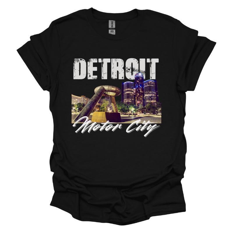 A black T-shirt featuring a bold Detroit-themed graphic. The design showcases a nighttime view of the city's skyline with the iconic Joe Louis Fist monument in the foreground. 'DETROIT' is displayed in distressed white capital letters at the top, while 'Motor City' is written in a sleek cursive font below. The design captures the energy and pride of Detroit.