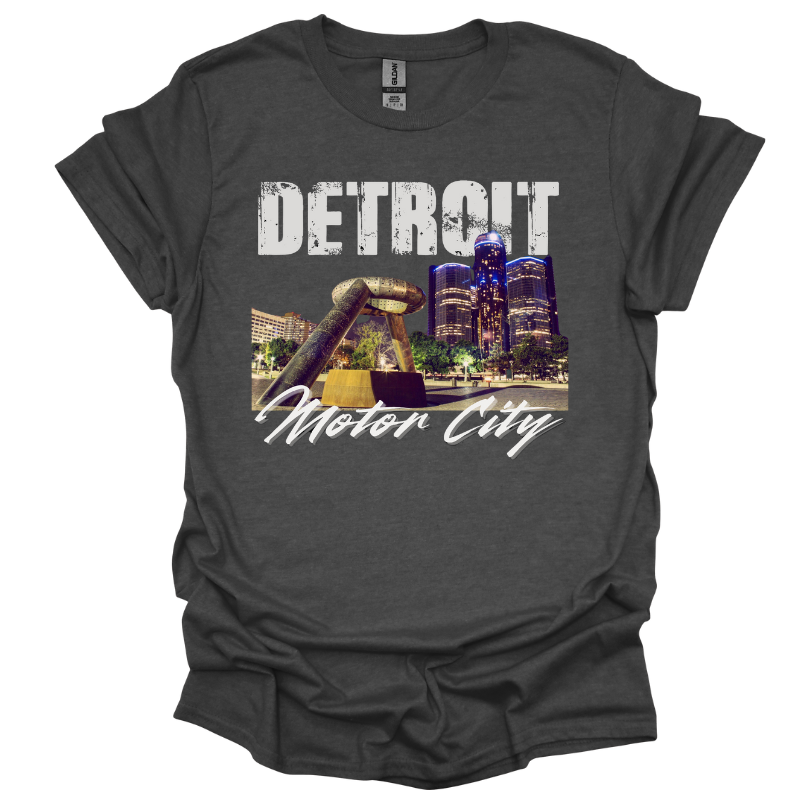 A gray T-shirt featuring a bold Detroit-themed graphic. The design showcases a nighttime view of the city's skyline with the iconic Joe Louis Fist monument in the foreground. 'DETROIT' is displayed in distressed white capital letters at the top, while 'Motor City' is written in a sleek cursive font below. The design captures the energy and pride of Detroit.