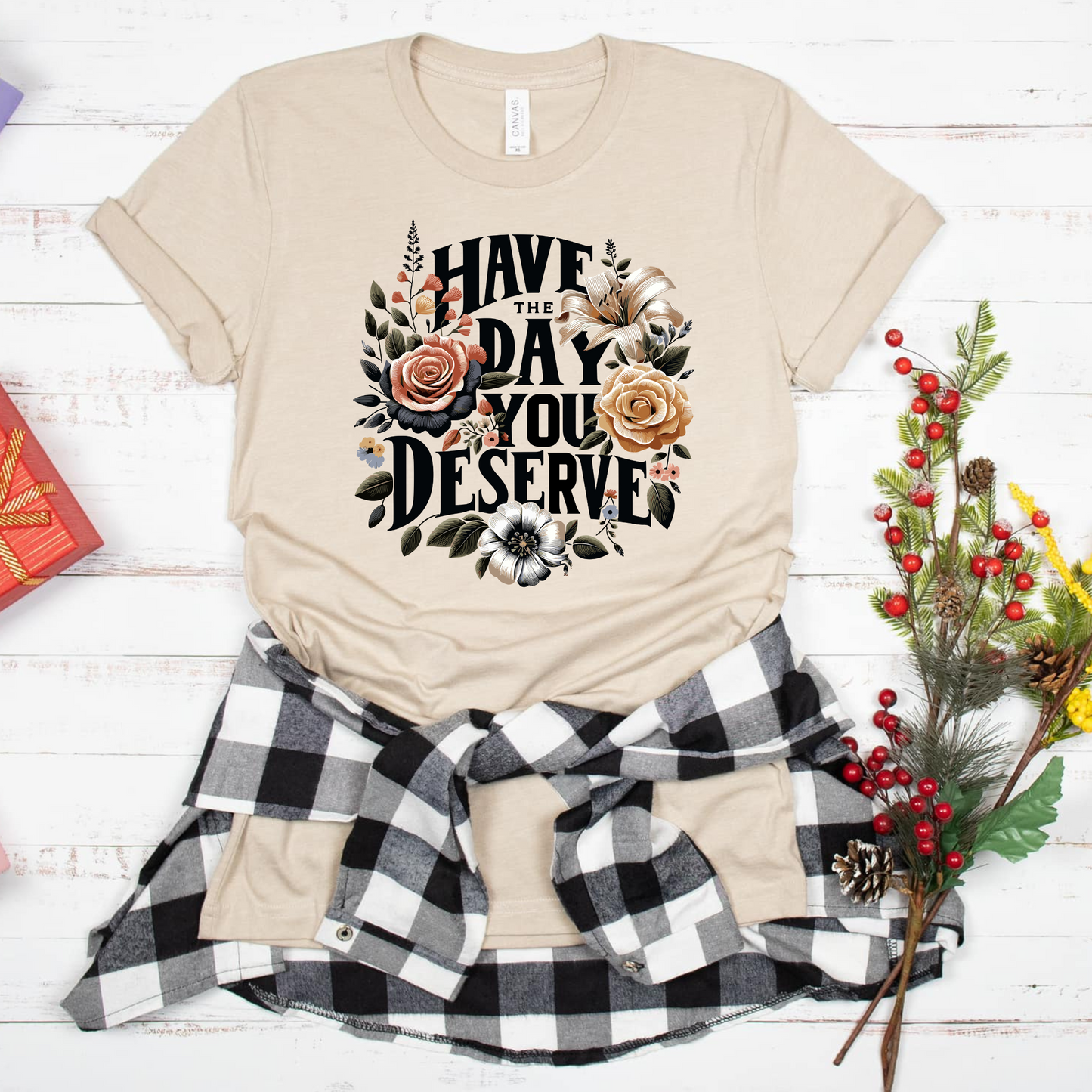 A beige T-shirt with the phrase 'Have the Day You Deserve' printed in bold, black lettering, surrounded by an intricate floral design featuring roses and other detailed flowers in warm, neutral tones.