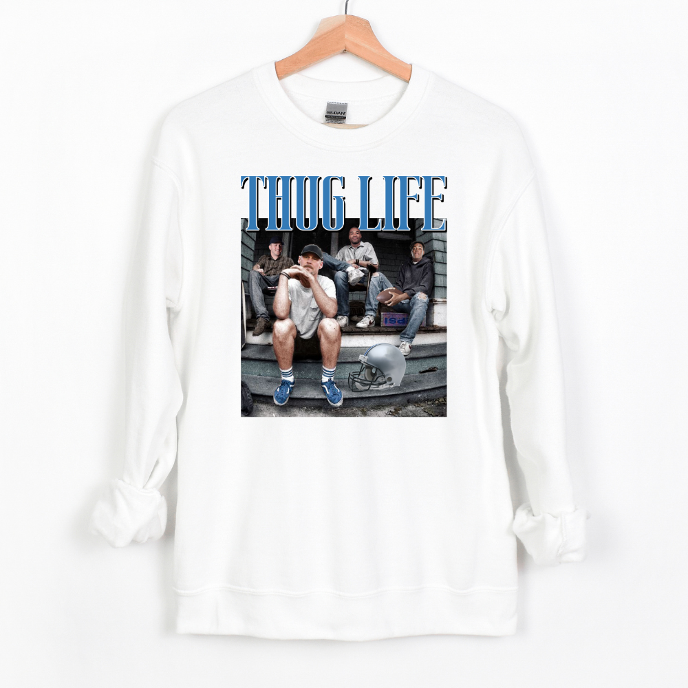 A white crewneck sweatshirt featuring Detroit football figures Jared Goff, Calvin Johnson (Megatron), Dan Campbell, and Jahmyr Gibbs sitting on a porch. Jared Goff is in the foreground wearing a cap and blue sneakers, with a football helmet placed on the ground nearby. The words 'THUG LIFE' are printed in bold, blue Old English-style font above the image. The design has a humorous, nostalgic, and gritty vibe, perfect for Detroit football fans.