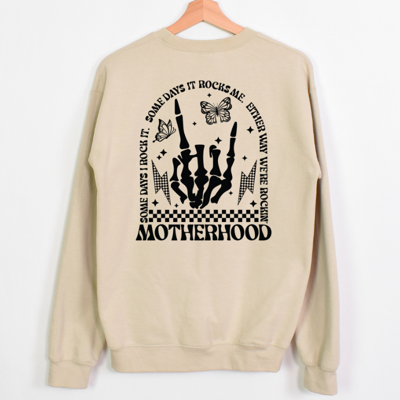 Rockin' Motherhood Crewneck is a natural color crewneck sweatshirt with a bold black graphic on the back. The design includes a skeleton hand making a rock sign, butterflies, lightning boltd, and a checkered pattern, with the phrase "some days I rock it. Some days it rocks me. Either way, we're rockin". Motherhood"