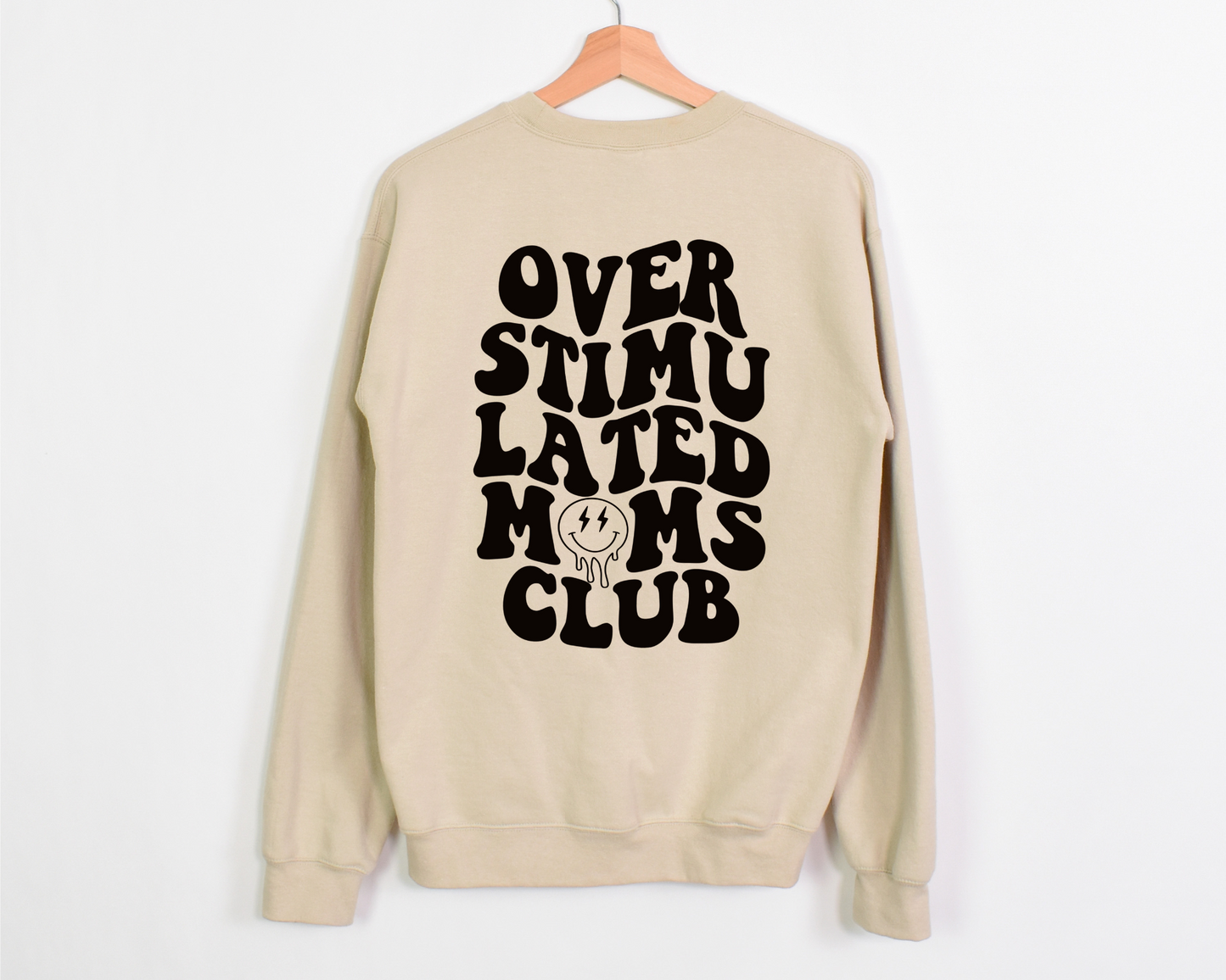 A natural colored crewneck featuring bold, wavy black text that reads 'Overstimulated Moms Club' on the back. The design includes a melting smiley face with lightning bolt eyes, capturing the chaotic and exhausted yet humorous side of motherhood.
