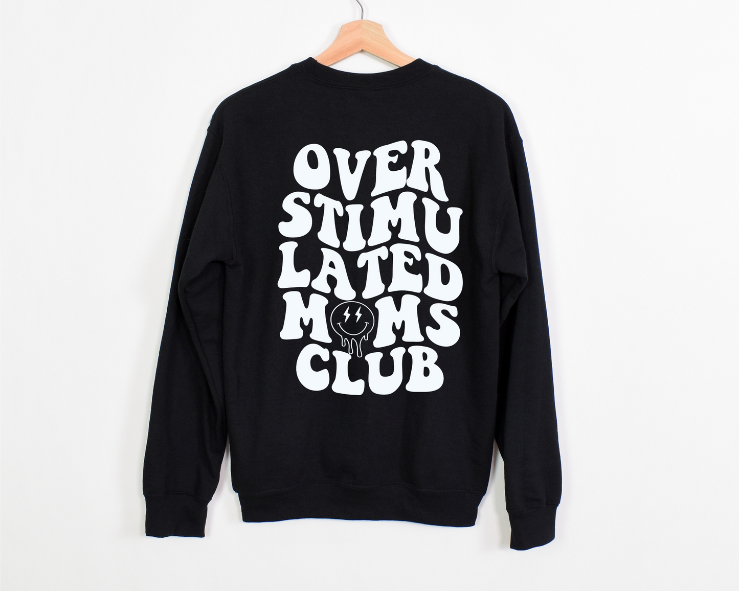 A black crewneck featuring bold, wavy white text that reads 'Overstimulated Moms Club' on the back. The design includes a melting smiley face with lightning bolt eyes, capturing the chaotic and exhausted yet humorous side of motherhood.