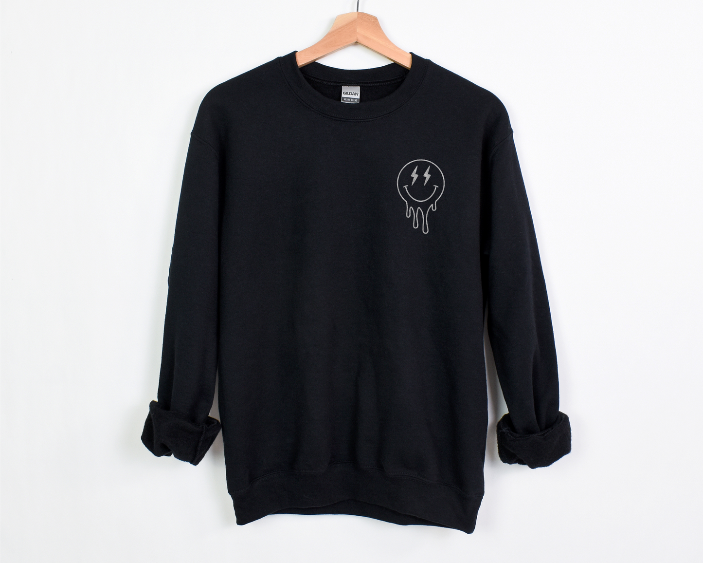 A black crewneck with a small white melting smiley face design on the left chest area. The smiley features lightning bolt eyes, giving it a playful yet overstimulated look.