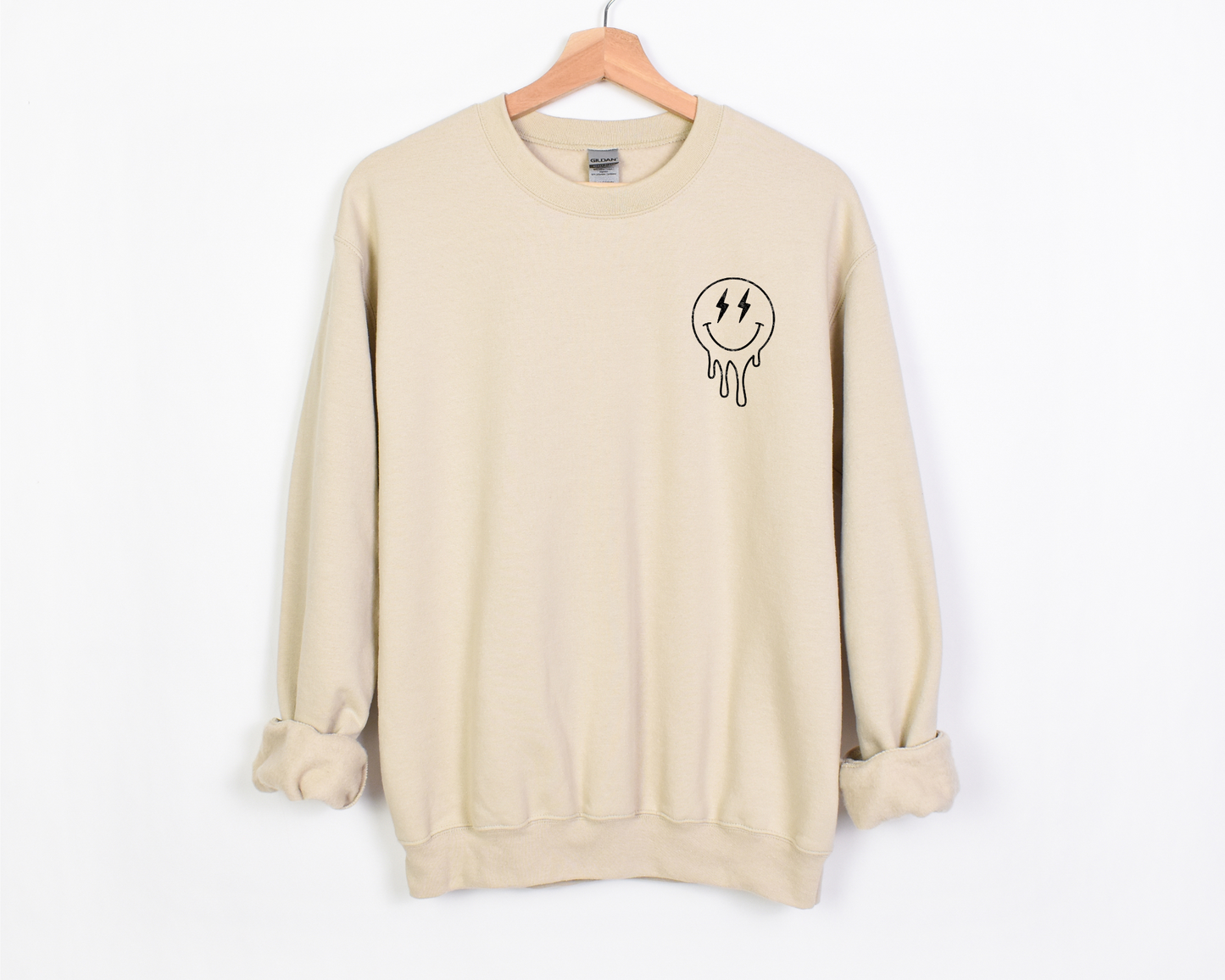A natural colored crewneck with a small black melting smiley face design on the left chest area. The smiley features lightning bolt eyes, giving it a playful yet overstimulated look.