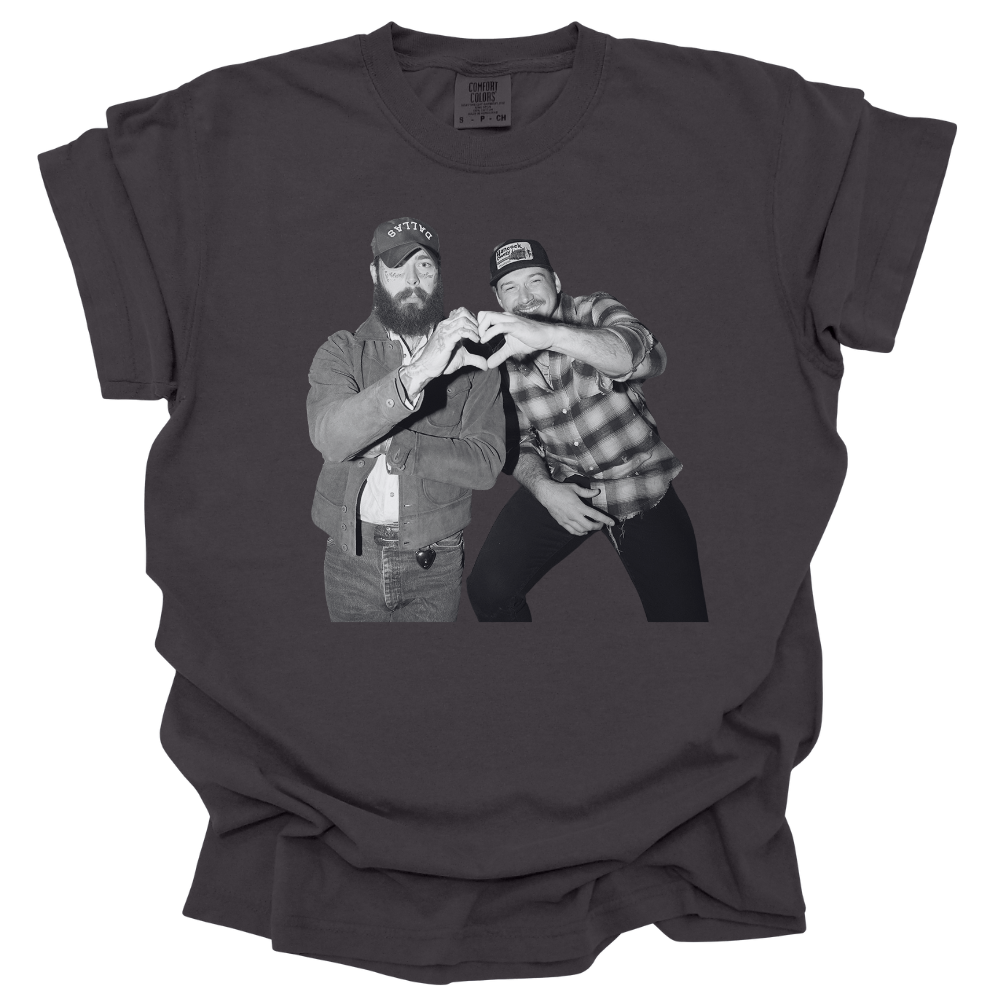 A graphite colored Comfort Colors T-shirt featuring a black and white image of Post Malone and Morgan Wallen in country-style outfits forming a heart shape with their hands. One wears a denim jacket and trucker hat, while the other sports a plaid shirt and baseball cap, both smiling and having a good time. The design has a vintage, laid-back country vibe.
