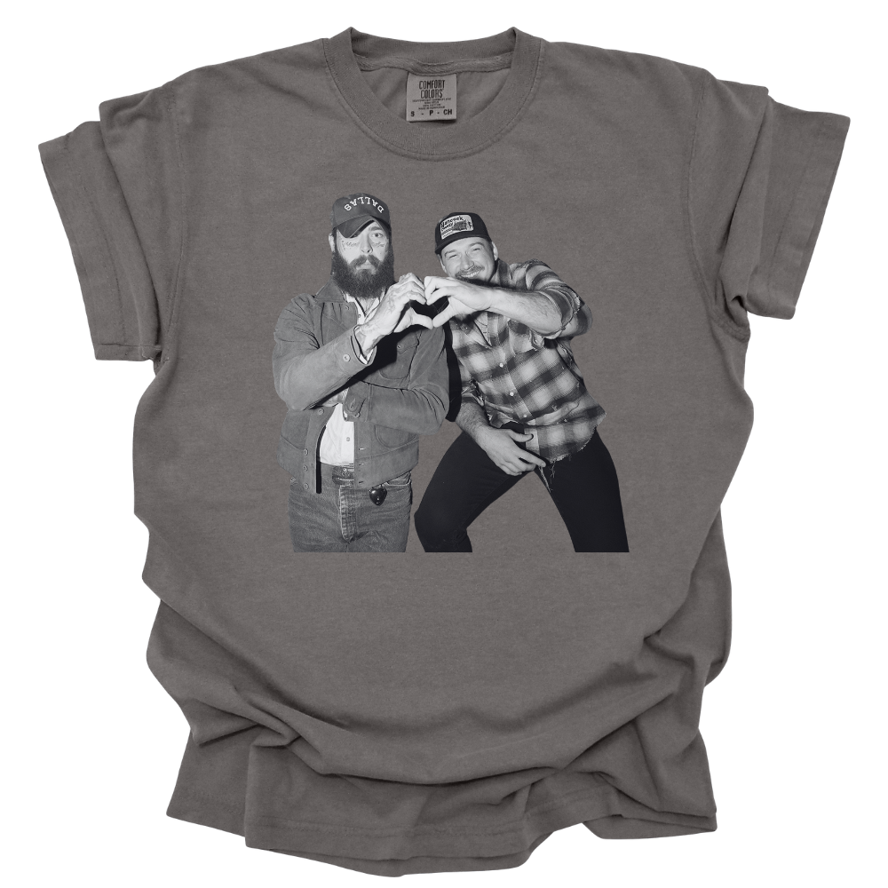A pepper colored Comfort Colors T-shirt featuring a black and white image of Post Malone and Morgan Wallen in country-style outfits forming a heart shape with their hands. One wears a denim jacket and trucker hat, while the other sports a plaid shirt and baseball cap, both smiling and having a good time. The design has a vintage, laid-back country vibe.