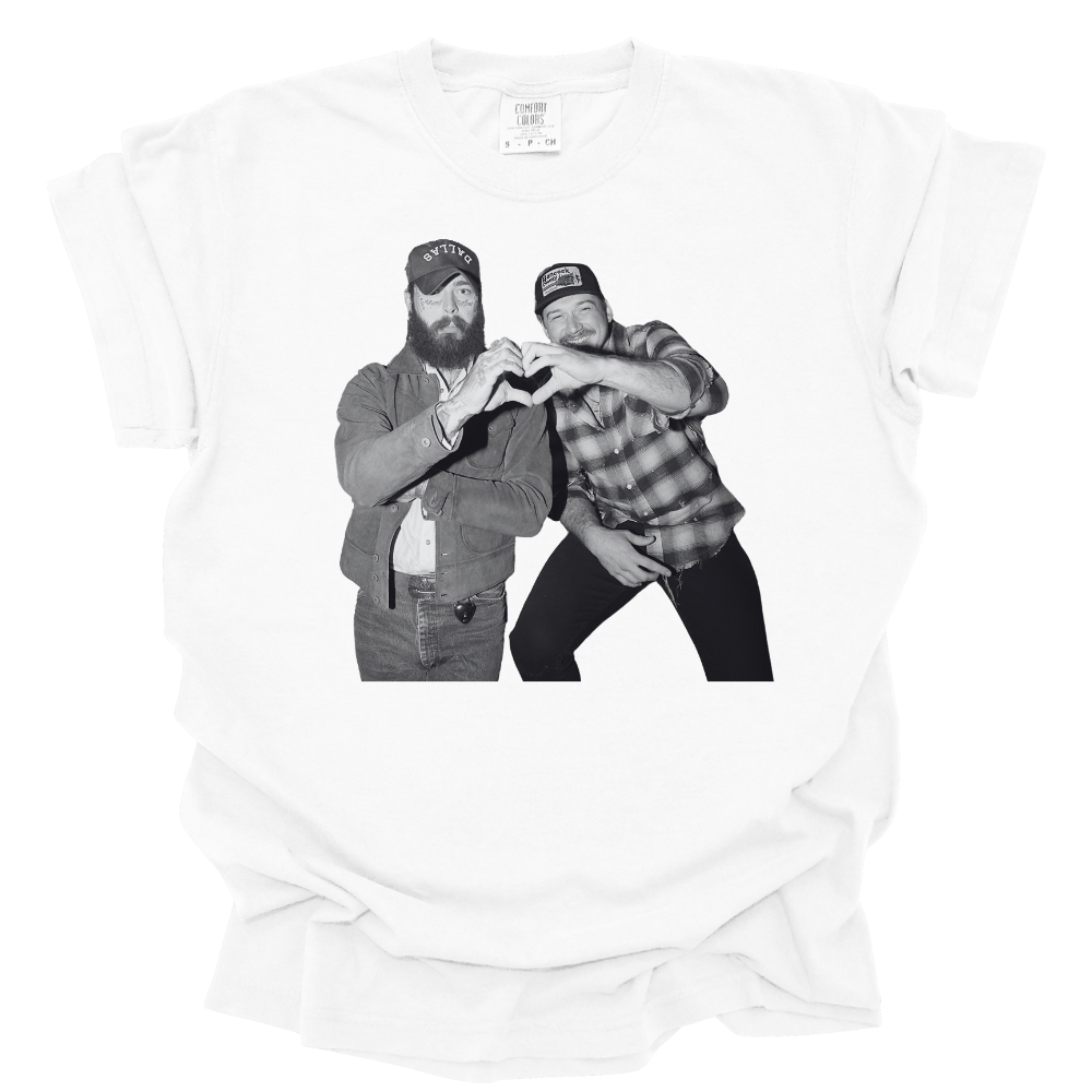 A white Comfort Colors T-shirt featuring a black and white image of Post Malone and Morgan Wallen in country-style outfits forming a heart shape with their hands. One wears a denim jacket and trucker hat, while the other sports a plaid shirt and baseball cap, both smiling and having a good time. The design has a vintage, laid-back country vibe.