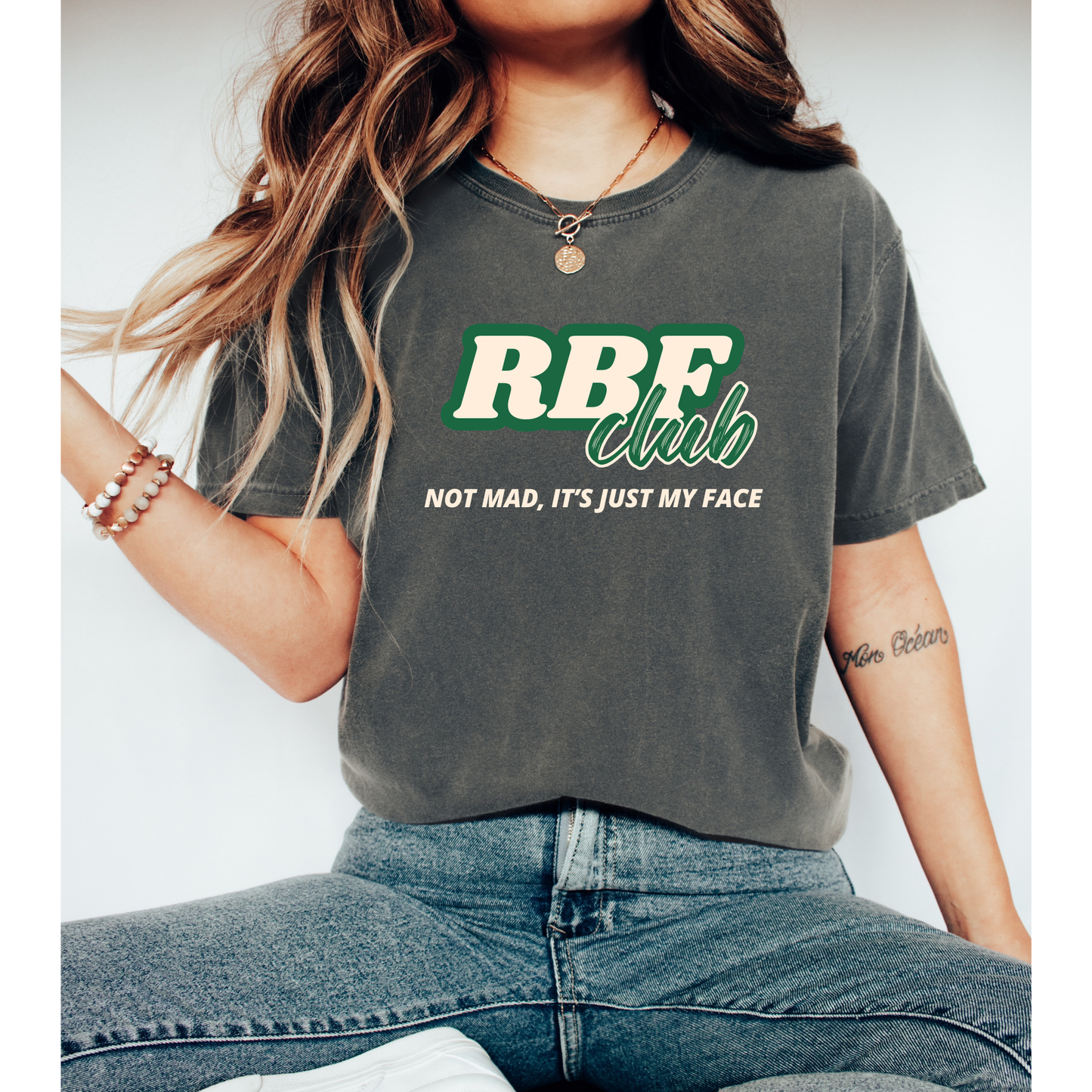 a vintage-washed, gray T-shirt with the words 'RBF Club' in bold green and white lettering across the chest. Below, the phrase 'Not Mad, It’s Just My Face' is printed in a smaller, italicized font. The relaxed-fit tee has a casual, worn-in look, styled with layered jewelry and rolled sleeves. The design gives off a playful and confident vibe