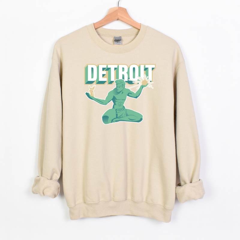 The Spirit of Detroit Crewneck is a sand colored crewneck sweatshirt featuring a stylized illustration of the Spirit of Detroit statue in green and teal tones. The design includes bold, retro-style "DETROIT" text with a 3D effect, giving it a modern yet classic city pride look