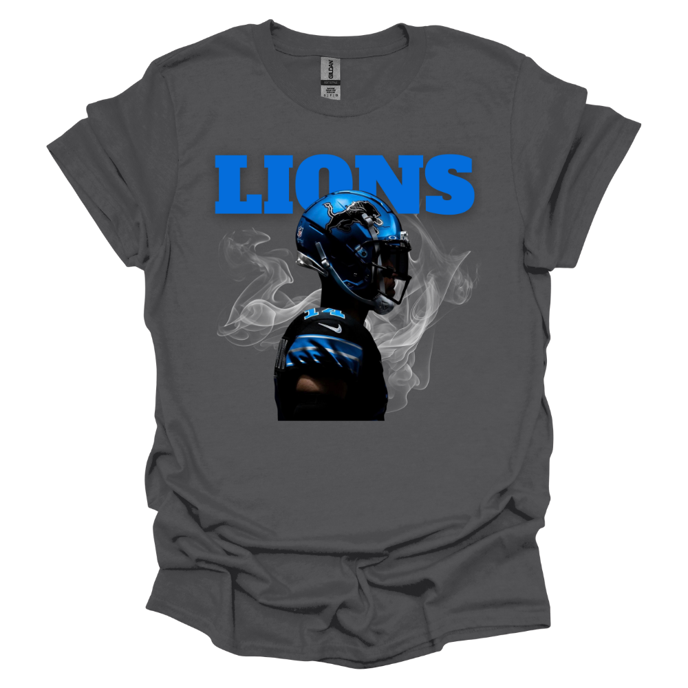 A gray t-shirt featuring a bold graphic of a football player wearing a blue Detroit helmet, surrounded by smoke. The word 'LIONS' is displayed in large, bright blue capital letters above the player. The high-contrast design gives the piece a dynamic and intense game-day feel, perfect for Detroit football fans