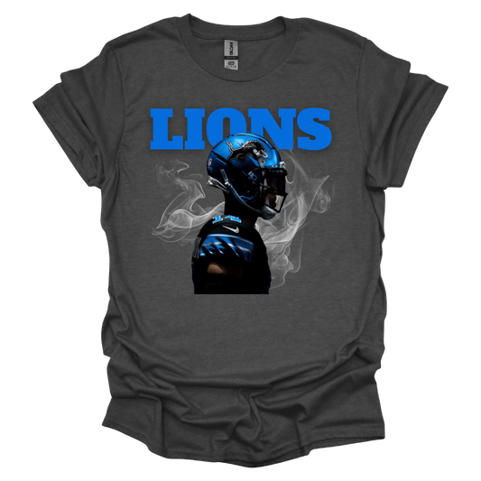 A dark gray t-shirt featuring a bold graphic of a football player wearing a blue Detroit helmet, surrounded by smoke. The word 'LIONS' is displayed in large, bright blue capital letters above the player. The high-contrast design gives the piece a dynamic and intense game-day feel, perfect for Detroit football fans
