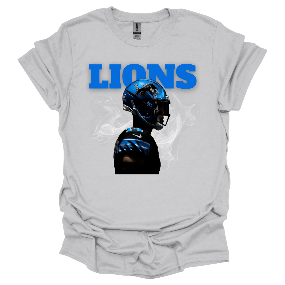 A light gray t-shirt featuring a bold graphic of a football player wearing a blue Detroit helmet, surrounded by smoke. The word 'LIONS' is displayed in large, bright blue capital letters above the player. The high-contrast design gives the piece a dynamic and intense game-day feel, perfect for Detroit football fans