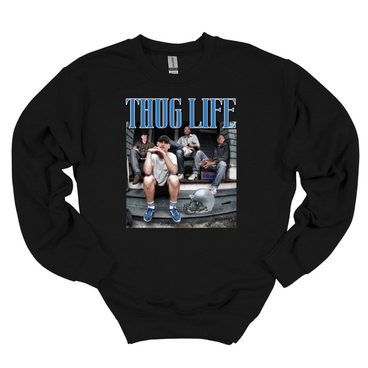 A black crewneck sweatshirt featuring Detroit football figures Jared Goff, Calvin Johnson (Megatron), Dan Campbell, and Jahmyr Gibbs sitting on a porch. Jared Goff is in the foreground wearing a cap and blue sneakers, with a football helmet placed on the ground nearby. The words 'THUG LIFE' are printed in bold, blue Old English-style font above the image. The design has a humorous, nostalgic, and gritty vibe, perfect for Detroit football fans.