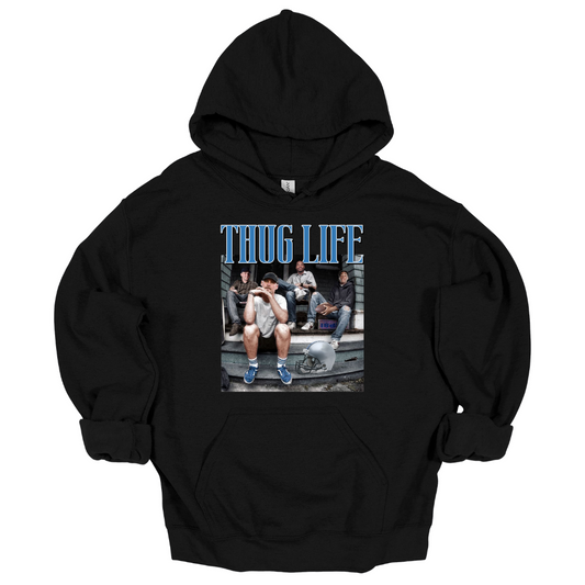 A black hoodie sweatshirt featuring Detroit football figures Jared Goff, Calvin Johnson (Megatron), Dan Campbell, and Jahmyr Gibbs sitting on a porch. Jared Goff is in the foreground wearing a cap and blue sneakers, with a football helmet placed on the ground nearby. The words 'THUG LIFE' are printed in bold, blue Old English-style font above the image. The design has a humorous, nostalgic, and gritty vibe, perfect for Detroit football fans.