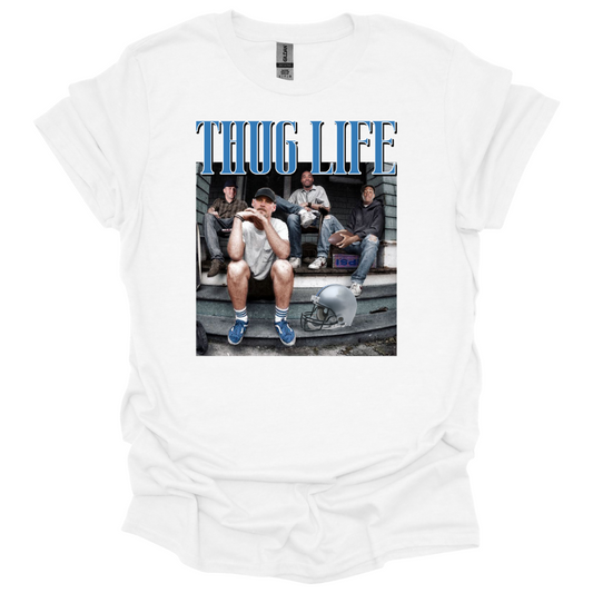 A white t-shirt sweatshirt featuring Detroit football figures Jared Goff, Calvin Johnson (Megatron), Dan Campbell, and Jahmyr Gibbs sitting on a porch. Jared Goff is in the foreground wearing a cap and blue sneakers, with a football helmet placed on the ground nearby. The words 'THUG LIFE' are printed in bold, blue Old English-style font above the image. The design has a humorous, nostalgic, and gritty vibe, perfect for Detroit football fans.